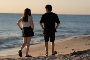 "Two people walking along a sandy beach at sunset, facing the sea and each other, with gentle waves rolling in the background and soft evening sunlight illuminating the shoreline."