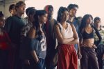 "A diverse group of young adults in sporty, casual clothing stand together indoors, appearing focused and attentive, possibly listening to someone or watching an event in a well-lit urban setting."
