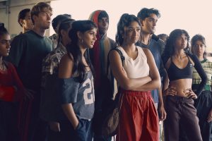 "A diverse group of young adults in sporty, casual clothing stand together indoors, appearing focused and attentive, possibly listening to someone or watching an event in a well-lit urban setting."