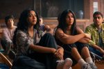 "A group of teenagers sitting on a gym floor, attentively listening, with two girls in the foreground and other students in the background in a school setting."