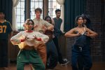 "A diverse group of young adults practicing dance in a studio with wooden floors, brick walls, and large windows. The two women in the front are focused on their hand movements, suggesting a choreographed routine."