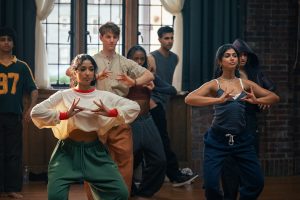 "A diverse group of young adults practicing dance in a studio with wooden floors, brick walls, and large windows. The two women in the front are focused on their hand movements, suggesting a choreographed routine."