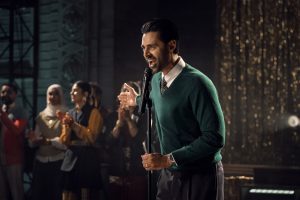 "Man in green sweater speaks into microphone on stage, with an audience of people clapping and watching in a warmly lit, festive indoor environment with golden decorations in the background."