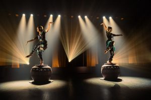 "Two performers in traditional attire balancing on large decorated drums on a stage, dramatic spotlights and beams illuminating them in a dark theater setting."