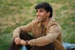"Smiling person with dark hair sitting outdoors on a blanket in a grassy area, wearing a tan jacket and brown pants, enjoying a relaxed moment in a park or natural setting."