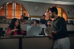"A young woman speaks to a man standing next to her in a college lecture hall; several students sit at desks with laptops, and sunlight filters through arched windows with wooden blinds in the background."