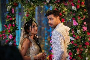 "A couple in traditional Indian attire stands under a vibrant floral arch at a decorated event, possibly a wedding or celebration, surrounded by colorful flowers and shimmering blue background lights."