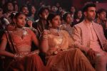 "Three people in ornate traditional South Asian attire sit in the front row of a crowded theater, watching a performance with serious expressions while the audience behind them reacts with interest and excitement."