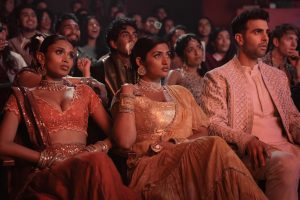 "Three people in ornate traditional South Asian attire sit in the front row of a crowded theater, watching a performance with serious expressions while the audience behind them reacts with interest and excitement."