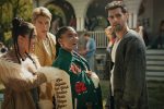 "Four young adults stand together outside at a social gathering, with a house and other people in the background, appearing to converse and enjoy a lively daytime event."