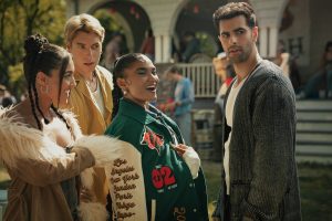 "Four young adults stand together outside at a social gathering, with a house and other people in the background, appearing to converse and enjoy a lively daytime event."