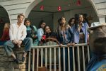 "Group of young adults on a porch at a casual gathering, dressed in trendy clothing, some holding drinks, with red cups hanging above and a bear statue wearing a shirt in the foreground, evoking a social, college party atmosphere."