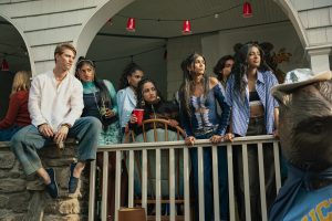 "Group of young adults on a porch at a casual gathering, dressed in trendy clothing, some holding drinks, with red cups hanging above and a bear statue wearing a shirt in the foreground, evoking a social, college party atmosphere."