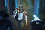 "A woman in a white corset, jeans, and a half-worn jacket stands confidently in a dimly lit room with blue lighting, beside a glowing crystal-like object on a pedestal, suggesting a dramatic or nightclub setting."