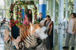 "Young woman in floral cart shaped like a tiger blowing smoke, surrounded by people in a bright modern building with large windows and greenery outside, some people dressed in colorful, traditional clothing."