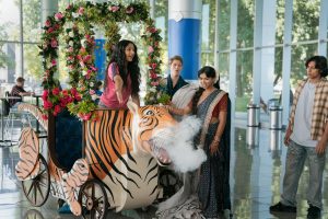 "Young woman in floral cart shaped like a tiger blowing smoke, surrounded by people in a bright modern building with large windows and greenery outside, some people dressed in colorful, traditional clothing."