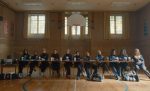 "A group of girls in athletic uniforms sit behind a long table in a gymnasium, with sports bags on the floor, under a basketball hoop and surrounded by tall windows and wall banners."