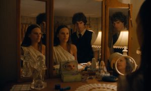 "A woman in a white dress sits at a vanity with a man standing behind her. Their reflections are seen in multiple mirrors, surrounded by various decorative items and soft lamp lighting in a warmly lit bedroom."
