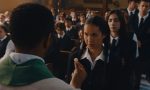 "A priest offers communion to a girl in a school uniform during a church service, with other students in uniforms seated and standing in the background inside a church setting."