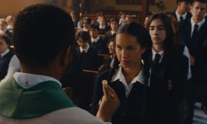 "A priest offers communion to a girl in a school uniform during a church service, with other students in uniforms seated and standing in the background inside a church setting."