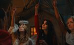 "Three women in costumes at a party, one dressed as an angel and another as a devil, raising their hands in a dimly lit room with rustic decor, suggesting a festive or Halloween atmosphere."