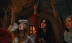 "Three women in costumes at a party, one dressed as an angel and another as a devil, raising their hands in a dimly lit room with rustic decor, suggesting a festive or Halloween atmosphere."