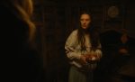 "Woman in a dimly lit wooden shed holding a wire basket with fresh eggs, wearing a light sweatshirt and looking at a person in the foreground, rustic interior with wooden plank walls and coop section in background."