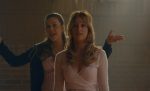 "Two young women stand in a warmly lit room with brick walls; one gestures and smiles energetically while the other looks ahead calmly, suggesting a lively conversation or instruction in a casual indoor setting."