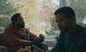 "Two men sitting indoors by a large window with trees outside, engaged in a serious conversation. One man wears a red jacket with a scarf, the other a dark sweater, both in a warmly lit, calm setting."