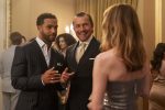 "Two men in suits and a woman in a metallic dress converse at an elegant indoor party, surrounded by other formally dressed guests in a well-lit, upscale setting with gold accents and curtains."