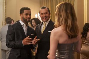 "Two men in suits and a woman in a metallic dress converse at an elegant indoor party, surrounded by other formally dressed guests in a well-lit, upscale setting with gold accents and curtains."