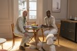 "Two men sitting at a round wooden table in a bright, elegant apartment with a window, fruit bowl, and modern decor. A white poodle on a leash sits on the floor beside them."