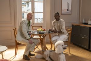 "Two men sitting at a round wooden table in a bright, elegant apartment with a window, fruit bowl, and modern decor. A white poodle on a leash sits on the floor beside them."