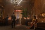 "A man in a suit stands in an elegant, wood-paneled lounge with a chandelier, while a woman in a gold dress sits on a couch; other people are visible in the background socializing at a formal event."