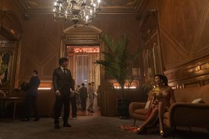"A man in a suit stands in an elegant, wood-paneled lounge with a chandelier, while a woman in a gold dress sits on a couch; other people are visible in the background socializing at a formal event."