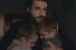 "A bearded man in a dimly lit room hugs two young boys close to his chest, providing comfort. All three have serious, emotional expressions, suggesting a moment of sadness or reassurance in a home setting."