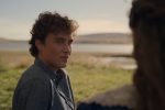 "Young man with curly hair outdoors in a grassy field near water, wearing a blue shirt, looking at a person with long hair whose back is to the camera, with hills and a calm sky in the background."
