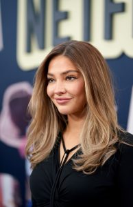 "Woman with long, wavy blonde hair in a black top posing on a red carpet at a premiere event, with a blurred Netflix logo and promotional background behind her."