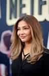 "Woman with long, wavy blonde hair in a black top posing on a red carpet at a premiere event, with a blurred Netflix logo and promotional background behind her."
