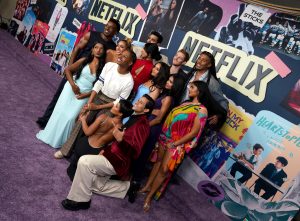 "Group of people posing together on a purple carpet at a Netflix event, with colorful posters and Netflix logos in the background, creating a lively and festive atmosphere."