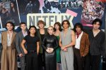 "A group of nine young adults pose together in front of a large Netflix sign at a premiere event, with colorful posters and graphics in the background, all dressed in stylish outfits."