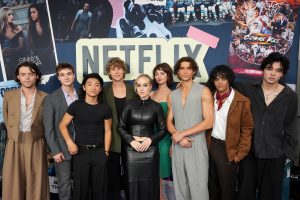 "A group of nine young adults pose together in front of a large Netflix sign at a premiere event, with colorful posters and graphics in the background, all dressed in stylish outfits."