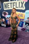 "Woman in a green patterned dress posing on a purple carpet at a Netflix event, in front of a collage wall with show posters and the Netflix logo."