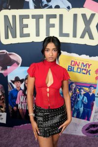 "Woman in a red top and black skirt posing at a Netflix promotional event, standing in front of a backdrop featuring posters for Netflix shows "On My Block" and "My Walter.""