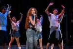 "A young woman with long red hair sings into a microphone on stage, surrounded by energetic dancers wearing casual clothes, all performing under stage lights in a dark theater setting."