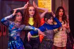 "Three young women in colorful, stylish outfits strike dramatic poses in a lively indoor setting, while a fourth woman in a white outfit stands behind them, watching with a neutral expression. The environment is vibrant and energetic."
