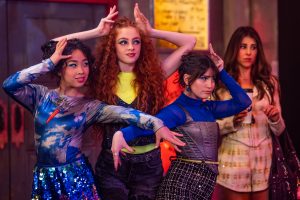 "Three young women in colorful, stylish outfits strike dramatic poses in a lively indoor setting, while a fourth woman in a white outfit stands behind them, watching with a neutral expression. The environment is vibrant and energetic."