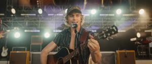 "Young male musician in a hat performs singing and playing guitar onstage at a concert, with stage lights, amplifiers, and instruments in the background."
