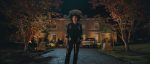 "Person in a black cowboy outfit stands on a driveway at night, with autumn trees on both sides and a large, well-lit house and parked cars in the background."