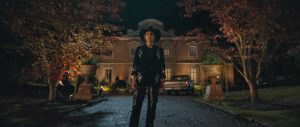 "Person in a black cowboy outfit stands on a driveway at night, with autumn trees on both sides and a large, well-lit house and parked cars in the background."